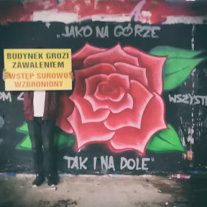 Zdjęcie autora strony na tle jego muralu z wizerunkiem czerwonej róży. Autor trzyma tabliczkę "Budynek grozi zawaleniem" na wysokości twarzy.