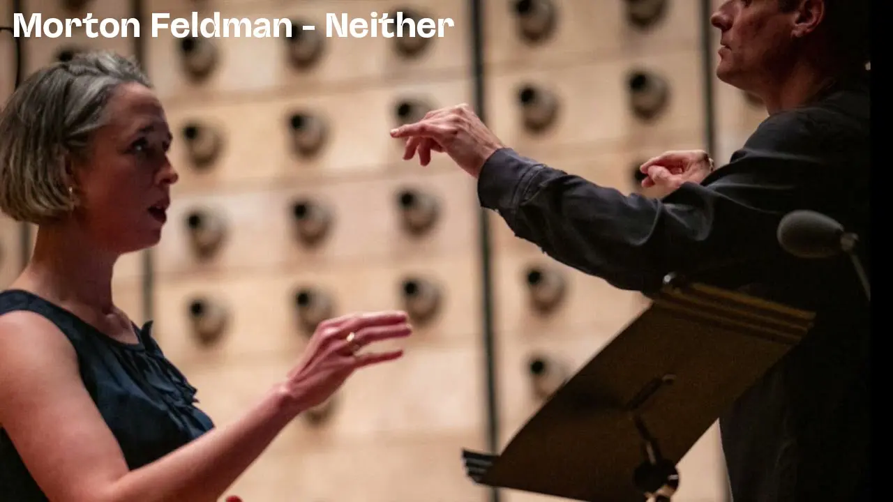 Two musicians conducting or performing music facing each other in a room with a wooden wall of circular acoustic panels, with text 'Morton Feldman - Neither'.