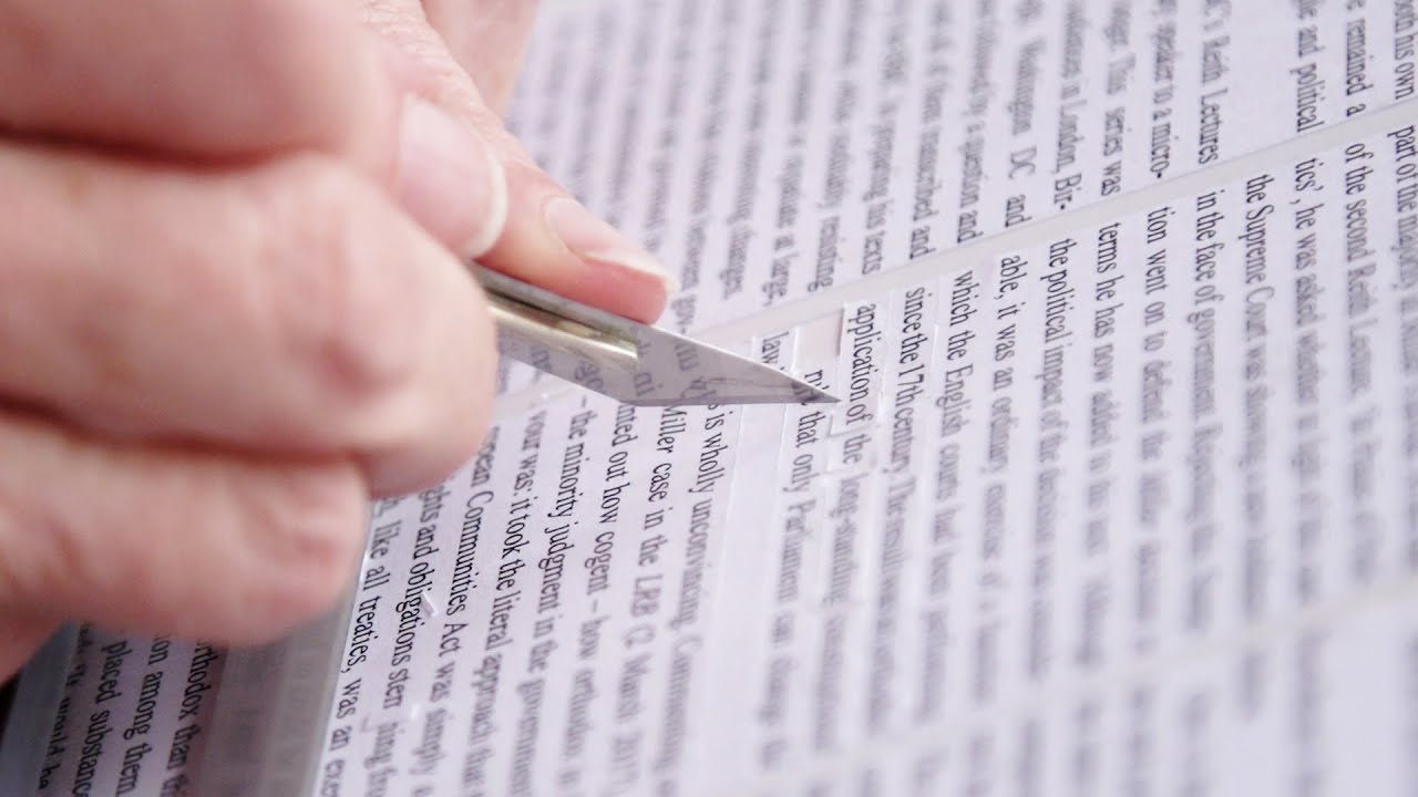 Close-up of a hand cutting a strip of text out of a newspaper with a craft knife.
